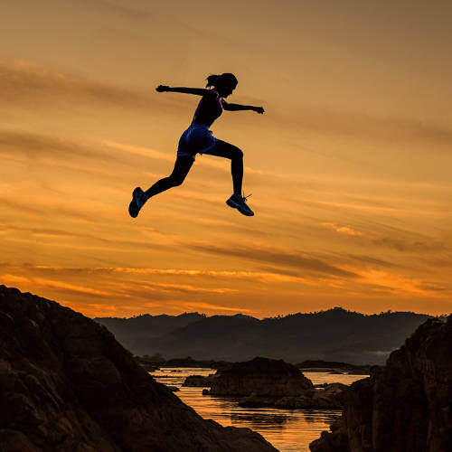A silhouette of a woman leaping across a wide gap between two cliffs at sunset, symbolizing resilience, mental toughness, and overcoming adversity.