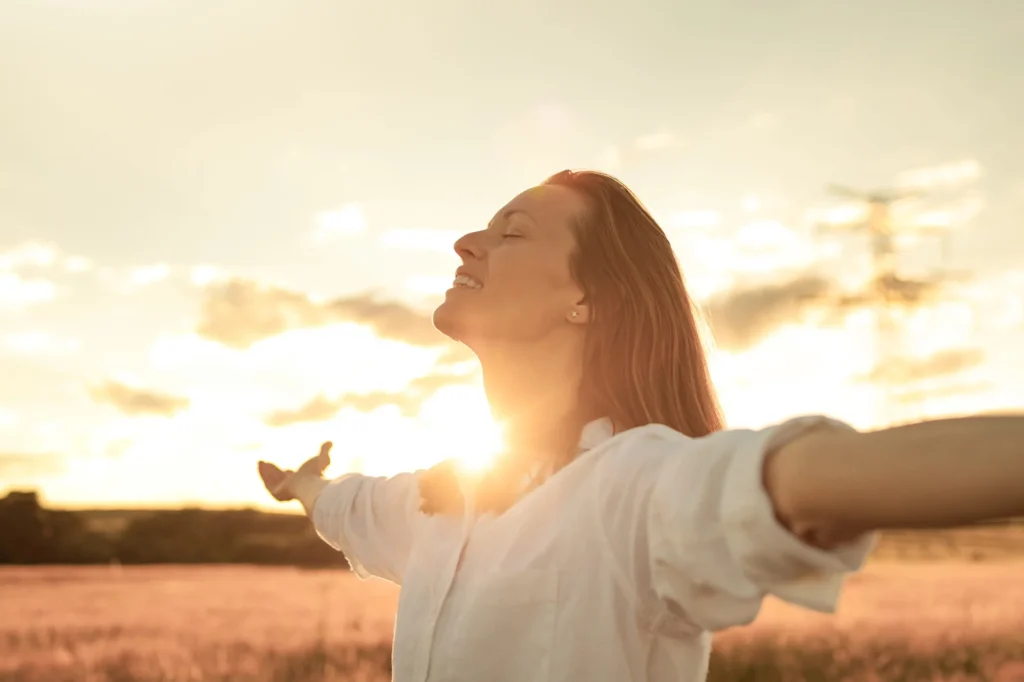 Woman with arms open and face turned toward the golden sunset or sunrise, celebrating freedom and emotional resilience in a field.