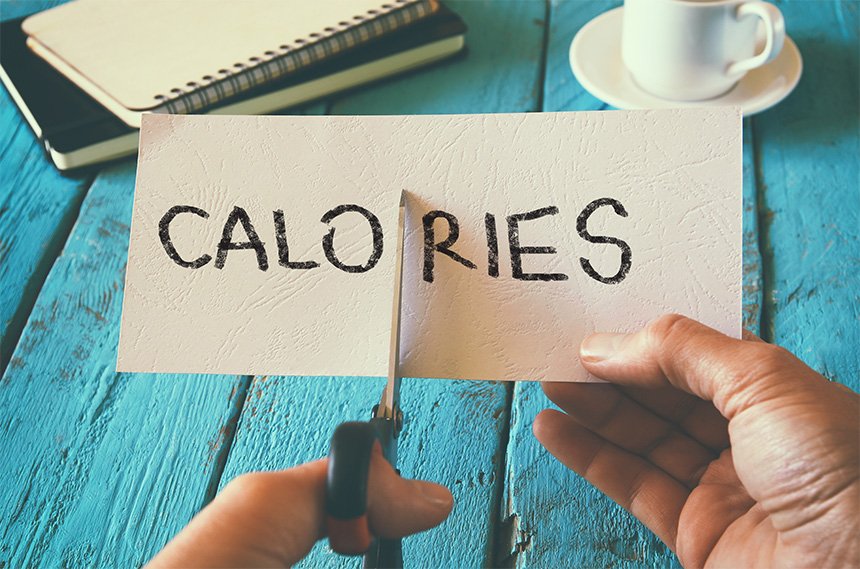A hand holding a pair of scissors cutting a piece of white paper with the word "CALORIES" written on it, visually representing the concept of cutting calories or achieving a calorie deficit. The background is a rustic blue wooden table with a notebook and a cup of coffee.